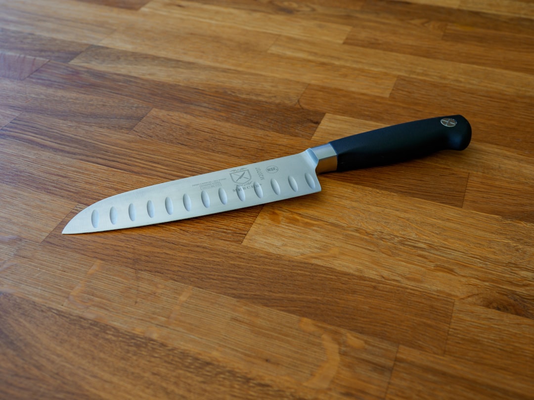 A close-up shot of a Santoku knife resting on a wooden cutting board surrounded by fresh vegetables,
